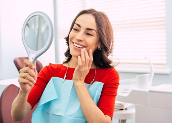 Woman with red-orange shirt touching teeth smiling at reflection in handheld mirror